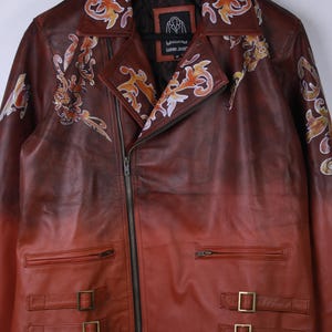 May include: A brown leather jacket with a gradient effect, transitioning from dark brown to red. The jacket features ornate floral designs in orange and white, a zipper closure, and two buckled straps at the waist.