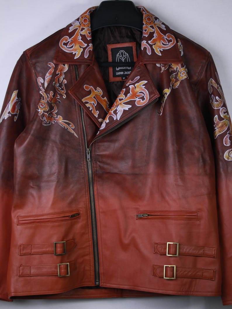 May include: A brown leather jacket with a gradient effect, transitioning from dark brown to red. The jacket features ornate floral designs in orange and white, a zipper closure, and two buckled straps at the waist.