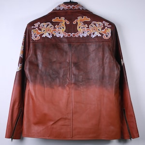 May include: A brown leather jacket with a gradient color effect, transitioning from dark brown to reddish-orange. The back features ornate, floral-like embroidery in shades of orange, yellow, and white. The jacket has a zipper on each sleeve.