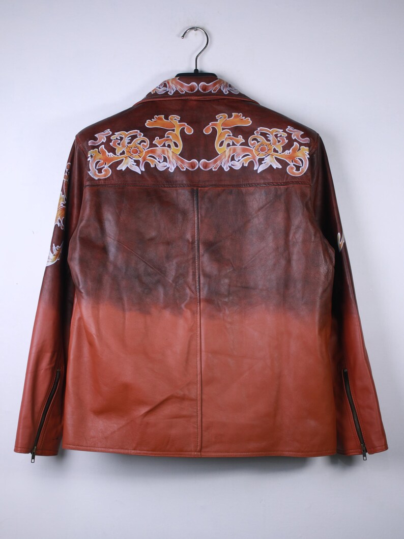 May include: A brown leather jacket with a gradient color effect, transitioning from dark brown to reddish-orange. The back features ornate, floral-like embroidery in shades of orange, yellow, and white. The jacket has a zipper on each sleeve.
