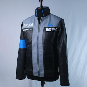 May include: A black and grey jacket with a high collar, featuring a blue armband on the right sleeve and a blue triangle on the left chest. The text RK 800, 13248317-51, and MADE IN DETROIT with the CyberLife logo are printed on the front panels.