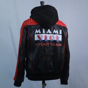 May include: A black and red jacket with a black hood, featuring the text MIAMI VICE STUNT TEAM in white and red on the back. The jacket has quilted red shoulder panels, long sleeves with red piping, and a black ribbed hem and sleeve cuffs.