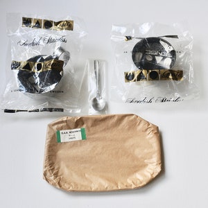 May include: Two clear plastic bags with black and gold labels that read "Swedish Stainless" and a small silver spoon. A brown paper tray with a green label that reads "GAS ROSTFRITT" and "Bollar" is in the foreground.