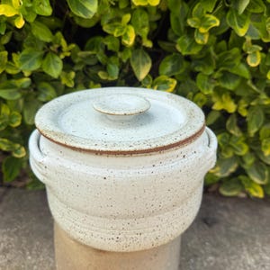 May include: A speckled white ceramic crock pot with a matching lid and small handles. The lid has a small knob. The crock pot is sitting on top of a larger, tan-colored ceramic container. Green foliage is in the background.