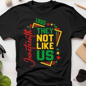 Puede incluir: Camiseta negra con el texto "1865 THEY NOT LIKE US" en amarillo y verde, con la palabra "Juneteenth" en rojo. La camiseta tiene cuello redondo y mangas cortas. Gafas de sol, un lápiz y otros accesorios también son visibles.