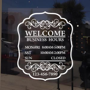 May include: A white decal on a glass door with the words "WELCOME BUSINESS HOURS" in a decorative frame. The hours are listed as Monday-Friday 8:00 AM-5:00 PM, Saturday 10:00 AM-2:00 PM, and Sunday closed. A phone number is also included.