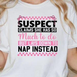 K&ouml;nnte beinhalten: Wei&szlig;es T-Shirt mit einer schwarzen und pinkfarbenen Grafik, auf der "SUSPECT CLAIMS SHE HAS SO MUCH TO DO BUT LAYS DOWN TO NAP INSTEAD" steht.