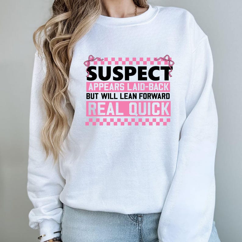 K&ouml;nnte beinhalten: Wei&szlig;er Pullover mit rosa Schriftzug "SUSPECT APPEARS LAID-BACK BUT WILL LEAN FORWARD REAL QUICK" mit rosa Schleifen auf beiden Seiten des Textes.