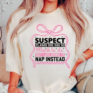 K&ouml;nnte beinhalten: Ein wei&szlig;es T-Shirt mit einer rosa Schleife und schwarzem Text, der "SUSPECT CLAIMS SHE HAS SO Much to do BUT LAYS DOWN TO NAP INSTEAD" lautet.