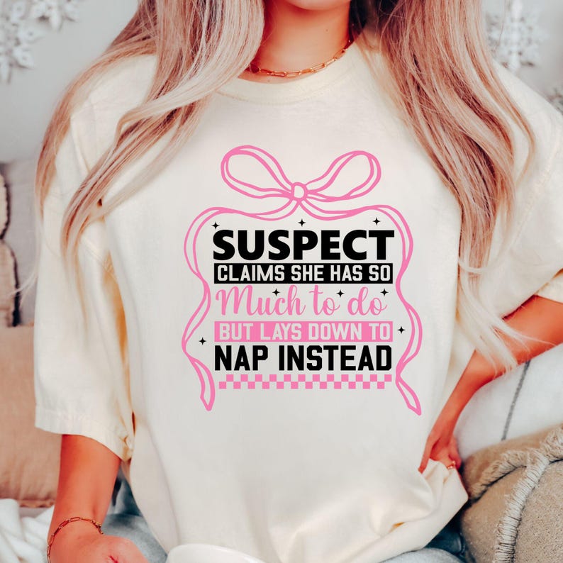 K&ouml;nnte beinhalten: Ein wei&szlig;es T-Shirt mit einer rosa Schleife und schwarzem Text, der "SUSPECT CLAIMS SHE HAS SO Much to do BUT LAYS DOWN TO NAP INSTEAD" lautet.