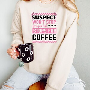 K&ouml;nnte beinhalten: Ein beiger Pullover mit dem Text "SUSPECT WON'T STOP for gas but *** STOPS FOR COFFEE" in rosa und schwarzer Schrift. Der Text ist von einem rosa-wei&szlig;en Karomuster umgeben.