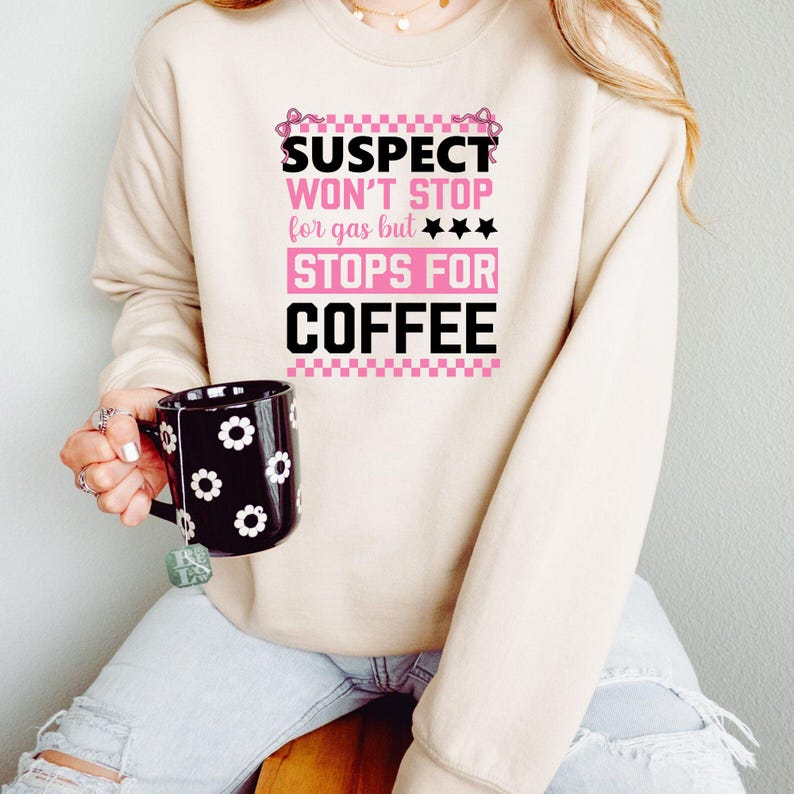 K&ouml;nnte beinhalten: Ein beiger Pullover mit dem Text "SUSPECT WON'T STOP for gas but *** STOPS FOR COFFEE" in rosa und schwarzer Schrift. Der Text ist von einem rosa-wei&szlig;en Karomuster umgeben.