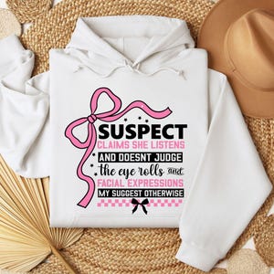 K&ouml;nnte beinhalten: Wei&szlig;er Hoodie mit einer rosa Schleife und schwarzem Text, der "SUSPECT CLAIMS SHE LISTENS AND DOESNT JUDGE the eye rolls and FACIAL EXPRESSIONS MY SUGGEST OTHERWISE" lautet.