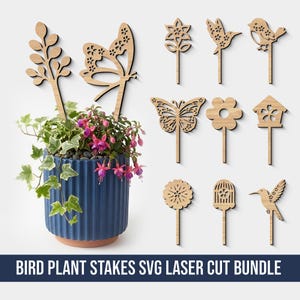 Op de afbeelding: Een verzameling lasergesneden houten plantenstokken met verschillende op de natuur geïnspireerde ontwerpen, waaronder vlinders, bloemen en vogels. De stokken worden getoond naast een blauwe keramische pot met een bloeiende plant. De tekst "BIRD PLANT STAKES SVG LASER CUT BUNDLE" staat onderaan.