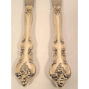 May include: Two silver-toned metal knife handles featuring an ornate, embossed floral and scrollwork pattern. The design includes intricate leaf motifs and small flower clusters extending from the base up the length of the handle.