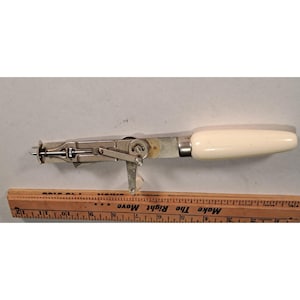 May include: Vintage can opener with a cream-colored handle and a silver-colored metal body. The can opener has a lever and a pointed tip. A wooden ruler is in the foreground.