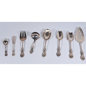 May include: A set of eight silver-toned metal serving utensils featuring ornate floral embossed handles. The collection includes a small spoon, a butter knife, a large fork, a ladle, a shell-shaped spoon, two serving spoons, and a cake server.