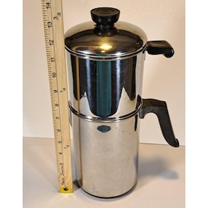 May include: A stainless steel coffee percolator with a black handle and lid knob. The percolator is approximately 13 inches tall, as measured by a wooden ruler. The coffee maker has a shiny, reflective surface.