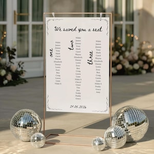 May include: A wedding seating chart with the text "We saved you a seat" and guest names. The chart is displayed on a copper-colored metal frame, with disco balls at the base. The date 24.05.2026 is printed at the bottom.