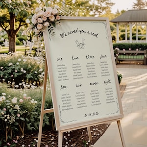 May include: A wedding seating chart on an easel, featuring a cream-colored background with black text. The sign reads "We saved you a seat" and lists names under numbers one through eight. Floral arrangements adorn the top.