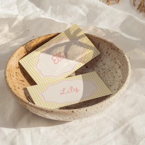 Peut inclure: Deux cartons de noms avec les noms "Ellie" et "Lily" en écriture rose, dans un bol en céramique mouchetée. Les cartes ont un fond rayé jaune et blanc avec une bordure rose festonnée.