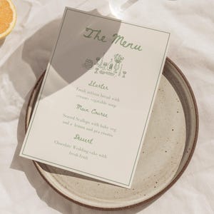 Peut inclure: Un menu avec le titre "The Menu" en écriture verte, listant une entrée, un plat principal et un dessert. Le menu est sur un fond beige, placé sur une assiette en céramique tachetée. Une tranche d'orange est visible dans le coin.