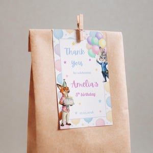 Pode incluir: Uma sacola de presente de papel marrom com uma etiqueta que diz "Obrigado por celebrar o 5&ordm; anivers&aacute;rio de Amelia". A etiqueta apresenta personagens de desenhos animados e bal&otilde;es coloridos. Um prendedor de madeira prende a etiqueta &agrave; sacola.