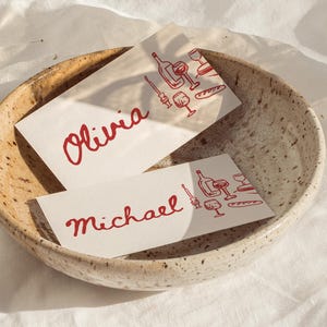 May include: Two place cards with the names "Olivia" and "Michael" in red script, each featuring a red illustration of wine bottles, glasses, and bread. The cards rest in a speckled, beige ceramic bowl.