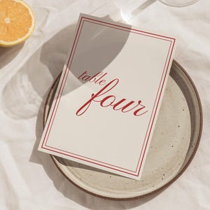 Puede incluir: Una tarjeta de mesa color crema con texto rojo "table four" dentro de un borde rojo. La tarjeta descansa sobre un plato de cerámica moteada. Una rodaja de limón y un vaso están en el fondo.