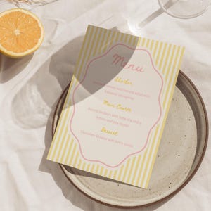 Peut inclure: Une carte de menu avec un fond à rayures jaunes et blanches et une bordure rose. Le menu répertorie une entrée, un plat principal et un dessert. Une demi-orange coupée et une assiette sont également visibles.