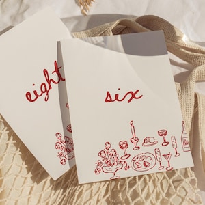 May include: Two white cards with red text and illustrations. One card reads "eight" and the other reads "six." The bottom of the "six" card features a red illustration of a table setting with a candle, wine glasses, and a bottle.