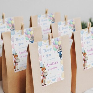 Pode incluir: Sacos de presente de papel castanho com etiquetas que dizem "Obrigado" e "5&ordm; anivers&aacute;rio da Amelia". As etiquetas apresentam personagens de desenhos animados e bal&otilde;es. Os sacos s&atilde;o fechados com molas de roupa de madeira.