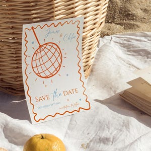 May include: A white save the date card with an orange border. The card features a red disco ball with a blue string hanging from it. The text on the card reads "James & Chloe Save the Date Saturday 24th May invitations to follow"
