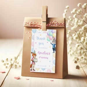 Pode incluir: Uma sacola de presente de papel marrom com uma etiqueta decorativa presa por um prendedor de madeira. A etiqueta apresenta personagens de desenhos animados, bal&otilde;es e o texto "Thank you for celebrating Amelia's 5th birthday 27.07.2018". A sacola &eacute; amarrada com barbante vermelho e branco.