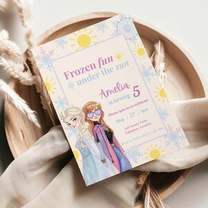 May include: A rectangular party invitation card featuring two animated characters with blonde and auburn hair, wearing blue and purple outfits. The text reads Frozen fun under the sun, Amelia is turning 5, May 27, 3pm, 168 Sunny Lane, Islington, London.
