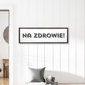 May include: A framed sign with the text "NA ZDROWIE!" in black lettering on a white background. The frame is dark brown wood.