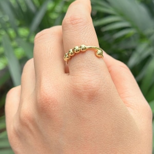 May include: A gold-colored ring featuring a series of small, round beads. The ring's design includes a curved section with the beads and a simple band. The ring is shown on a finger against a blurred green background.