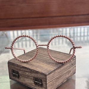 Könnte beinhalten: Eine runde Brille aus gedrehtem Kupferdraht. Die Fassung weist eine durchgehende seilartige Textur an den kreisförmigen Rändern, dem Steg und den Bügeln auf. Das metallische Kupfermaterial hat einen warmen, rotbraunen Farbton.