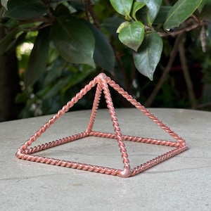 May include: A pyramid-shaped sculpture constructed from twisted copper wire. The structure features a square base and four angled sides that meet at a single apex point, with all edges formed by a continuous, spiraled metallic copper wire design.