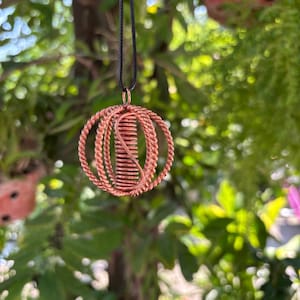 Puede incluir: Un colgante de alambre de cobre con un cilindro central enrollado rodeado por una jaula esférica de hebras de alambre trenzado. El colgante cuelga de un cordón negro fino y muestra un acabado metálico marrón rojizo con detalles de alambre.