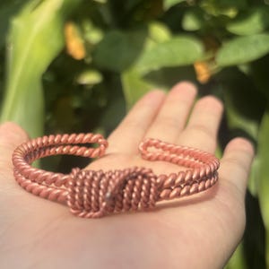 May include: A copper-colored cuff bracelet featuring a double-strand twisted wire design. The center of the bracelet is adorned with a prominent, rectangular knot detail created from the same twisted wire, forming an open-ended circular shape.