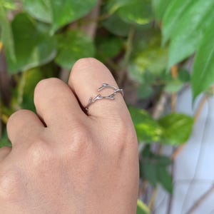 May include: A silver-colored ring in the shape of a delicate branch with small twigs. The ring is worn on a finger, showcasing its intricate design against a blurred green background.