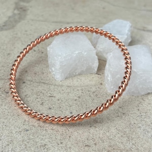 May include: A circular bangle bracelet crafted from a single continuous strand of polished copper metal. The bracelet features a consistent twisted rope texture throughout its entire circumference, creating a repeating spiral pattern in a warm metallic tone.