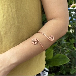 Può includere: Un bracciale da braccio in metallo color rame caratterizzato da un semplice design aperto con due terminali a spirale circolare alle estremità. La fascia simile a un filo avvolge l'avambraccio, con finitura metallica e struttura curva.