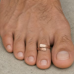 May include: A copper-toned toe ring featuring an open-ended, adjustable design with a textured, hammered finish. The band wraps around the second toe, showcasing a metallic sheen and a distinct, rugged surface pattern across its entire outer face.