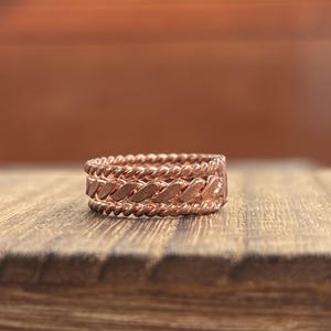 May include: A copper-colored ring featuring a central band with a diagonal braided texture, flanked by two thinner bands with a twisted rope design. The metal has a warm, metallic finish with visible ridges and grooves across the entire surface.
