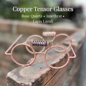 Könnte beinhalten: Copper Tensor Glasses mit Rosenquarz, Amethyst und Lapislazuli. Die Brille besteht aus gedrehtem Kupferdraht, wobei ein blauer Lapislazuli-Kristallstab über dem Steg zwischen den beiden kreisförmigen Drahtrahmen positioniert ist.