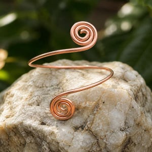 May include: A copper wire arm cuff featuring a continuous band that curves into two distinct spiral ends. The metal has a polished, warm reddish-brown metallic finish, and the design consists of a single piece of coiled wire forming a circular shape.