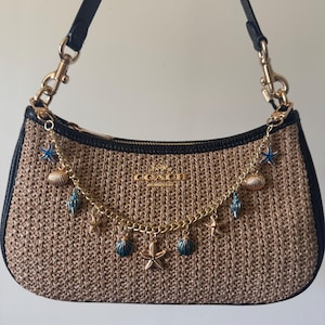 May include: A brown straw Coach handbag with a black leather trim and a gold chain strap with charms of seashells and starfish.