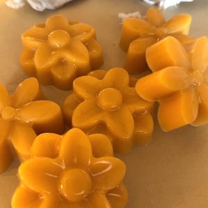 May include: Close-up of several flower-shaped, golden-yellow soaps. The soaps have a glossy finish and are arranged on a light brown surface. Small, off-white drawstring bags are visible in the background.